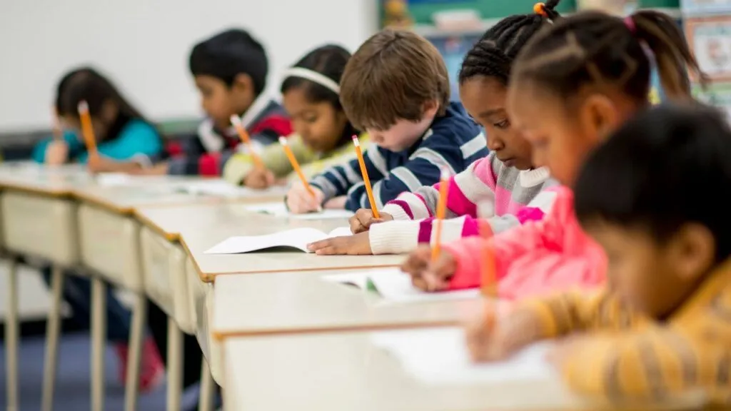 Children sitting at desks arranged in a semicircle, writing in their notebooks