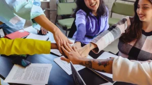 Young adults sitting around a table, joining their hands in the center.